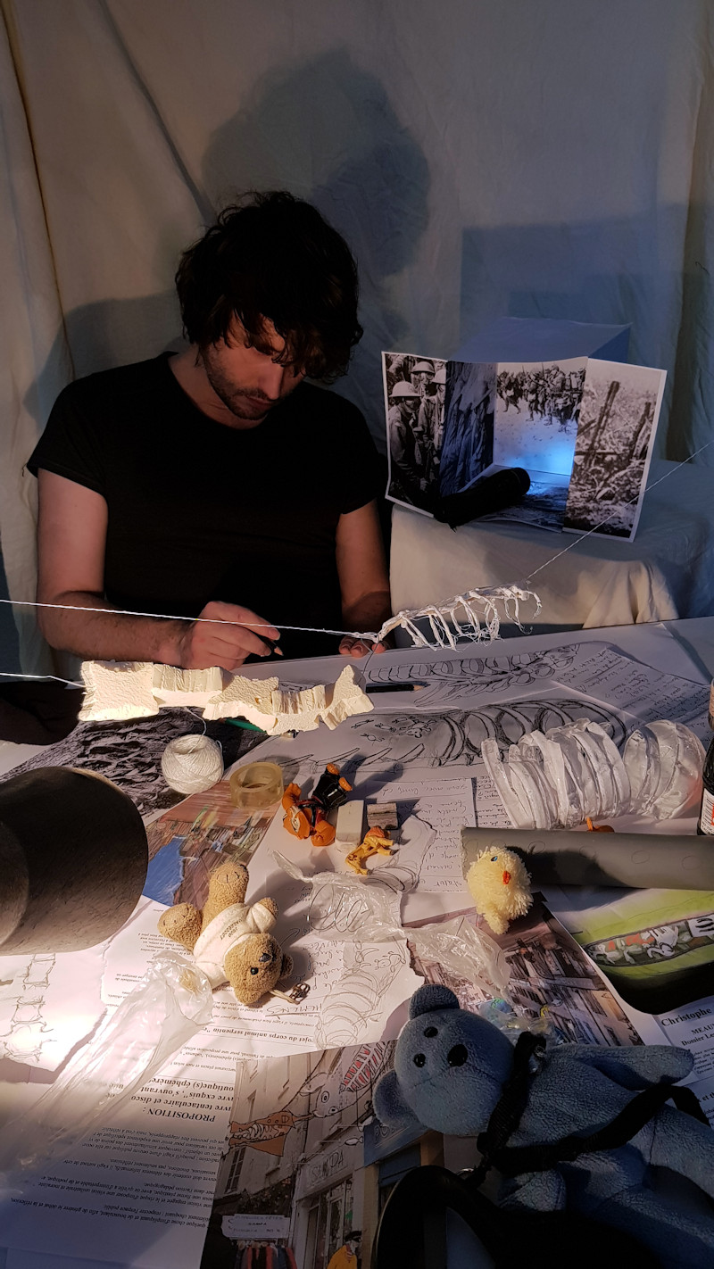 Dans le cadre feutré de son atelier, Christophe Alzetto prépare sa nouvelle grande exposition. Devant lui, toutes sortes d'objets comme autant d'indices de son projet : Maquettes suspendues, nounours, ébauches de structures squelettiques, croquis et notes de travail.